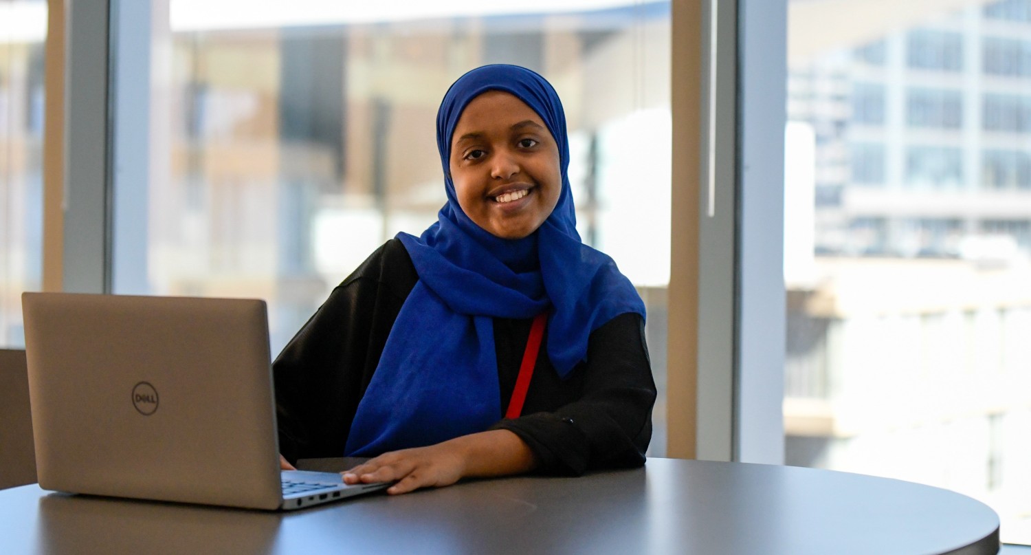 A young woman wearing a hijab is smiling at the camera, using a laptop.