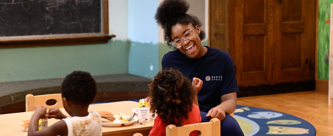 Intern is sitting at a preschool table. She is smiling and engaging with a toddler during a meal, while another toddler is also at the table.