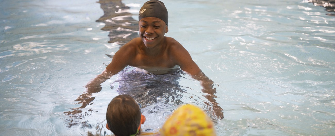 Intern is in a swimming pool wearing a black swim cap, smiling. He is facing a young swim student who is coming toward them.
