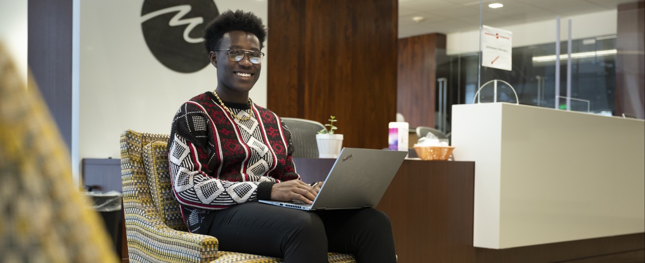 An intern is wearing a sweater and sitting with an open laptop in a lobby chair.