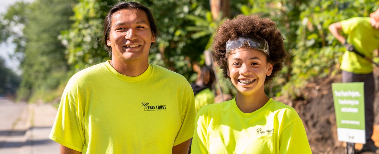 Two interns are wearing bright green T-shirts. They are standing outside with trees in the background.